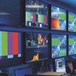 A monitoring room with a long desk with monitors on. Screens also line the walls showing a sports game